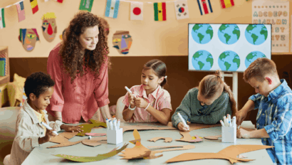 A teacher supervises four children working on a craft project at a classroom table decorated with international flags and cut-out materials.