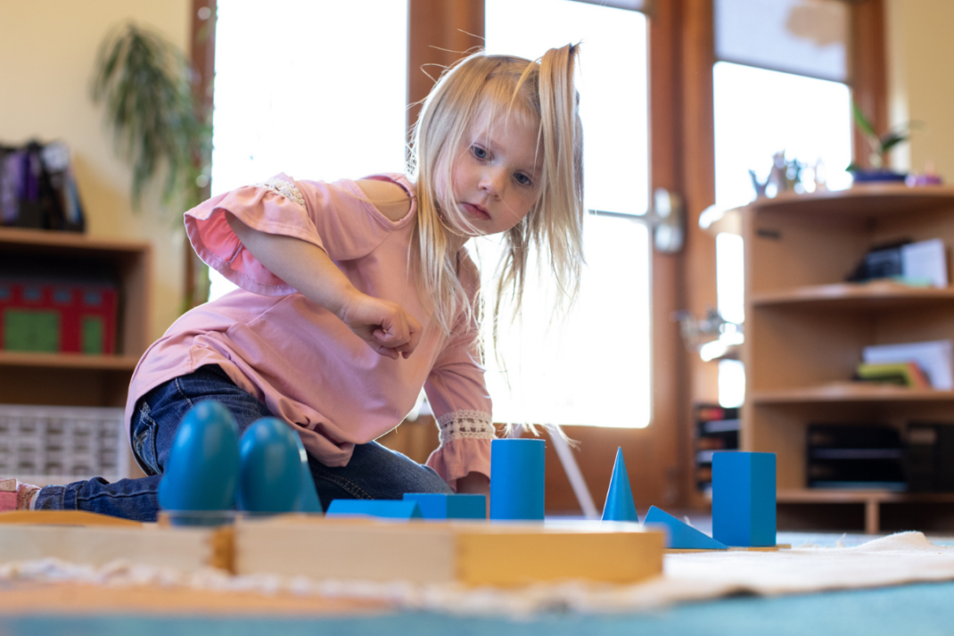 Young girl in a pink shirt sits on the floor indoors, engaging with blue geometric shapes on a mat in a brightly lit room.
