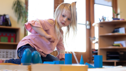Young girl in a pink shirt sits on the floor indoors, engaging with blue geometric shapes on a mat in a brightly lit room.